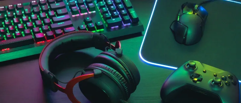 A close-up shot of gaming peripherals including a mechanical keyboard with glowing RGB keys, a headset, a gaming mouse on a mousepad, and a black game controller, all lit with green and magenta light.